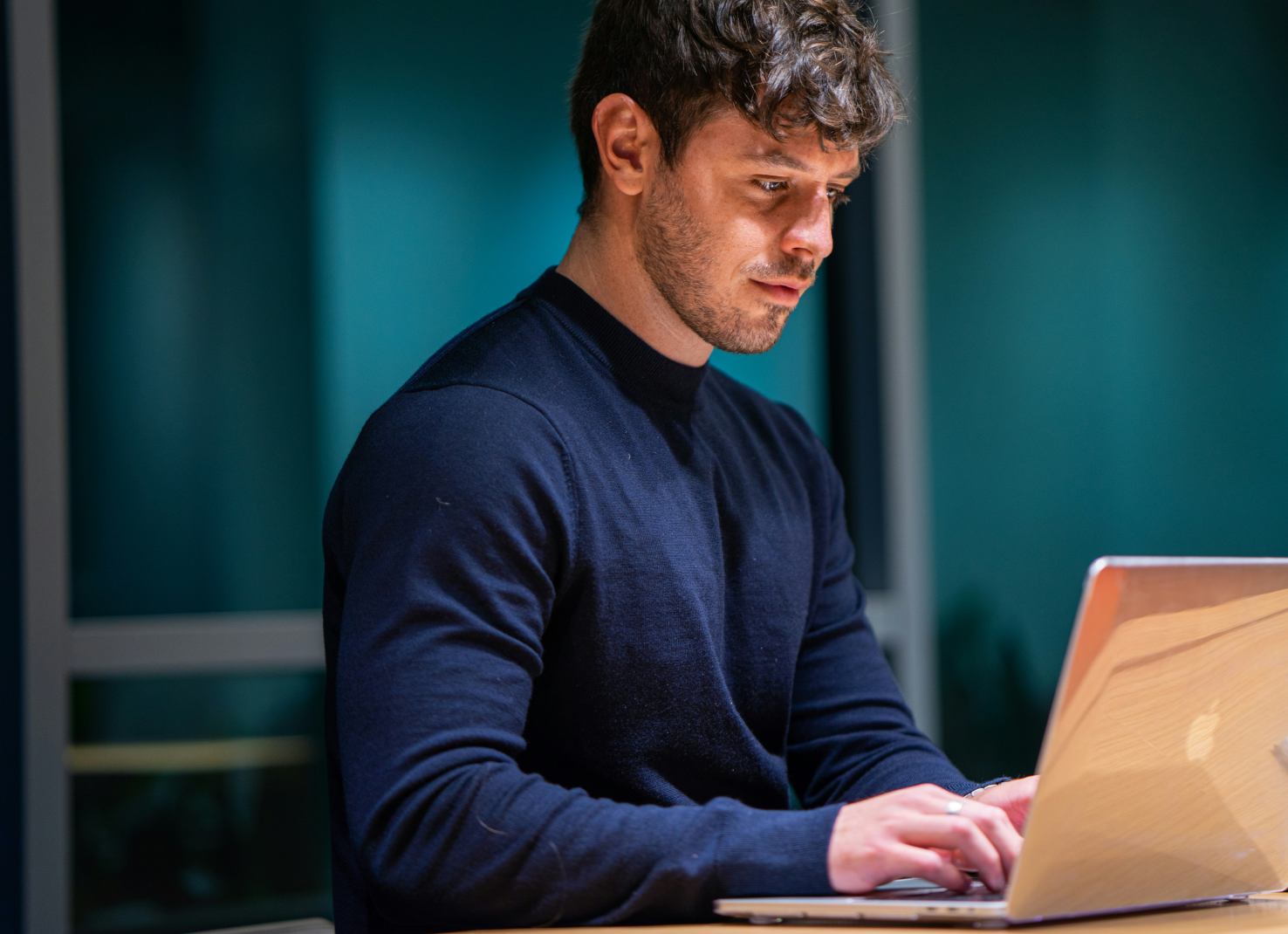 Young man in a navy sweater working intently on a laptop at night in a modern office with dim lighting