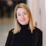 Valeria Corti, Jetico Marketing Manager and News Writer, posing in a modern office hallway, wearing a black blazer and turtleneck, with a confident and approachable expression.