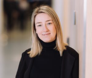 Valeria Corti, Jetico Marketing Manager and News Writer, posing in a modern office hallway, wearing a black blazer and turtleneck, with a confident and approachable expression.